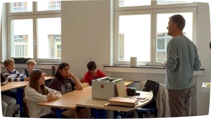 Lehrkraft steht vor einer Klasse und spricht mit Schülerinnen und Schülern im Unterricht.
