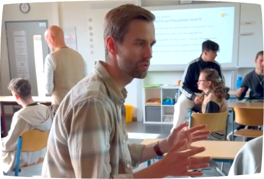 Lehrkraft leitet eine Unterrichtsdiskussion, während Präsentationsinhalte im Hintergrund angezeigt werden.